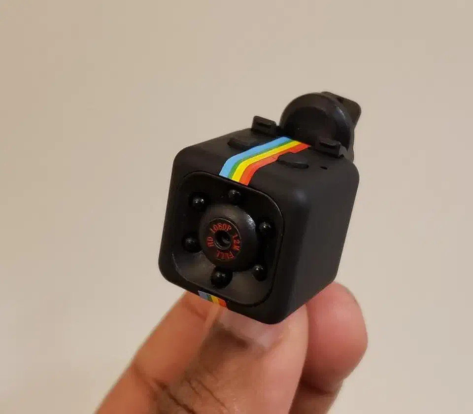 SQ11 mini camera held between fingers showing the front lens, infrared LEDs, and rainbow stripe